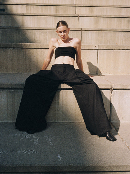 Person wearing a black strapless top and black wide-leg pleated pants sitting on concrete steps 