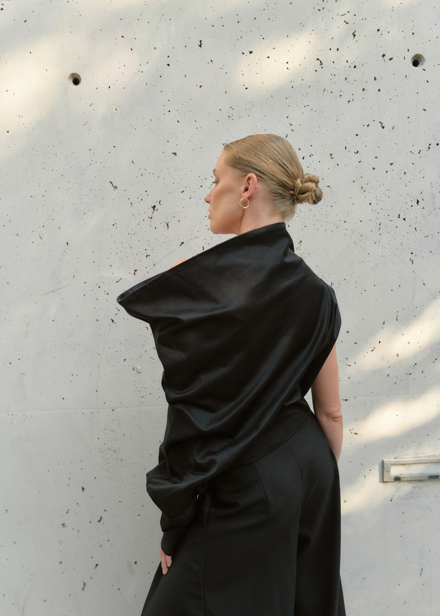 Back view of a sculptural black asymmetric top with a high draped collar and exposed shoulder
